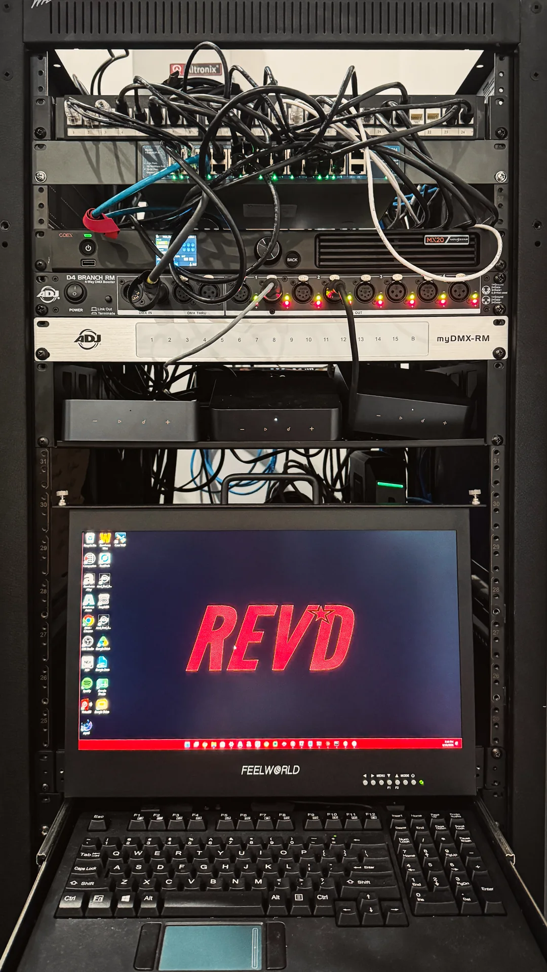 An equipment rack with a patch panel and network switch, LED processor, four-way DMX booster and myDMX interface, a shelf of media players, and a pull-out console whose rack monitor shows the REVD desktop.