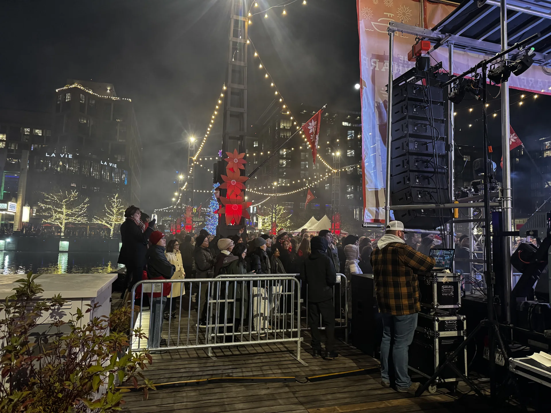 Crowd behind barricade at the pier stage, with line array, string lights and the holiday tree