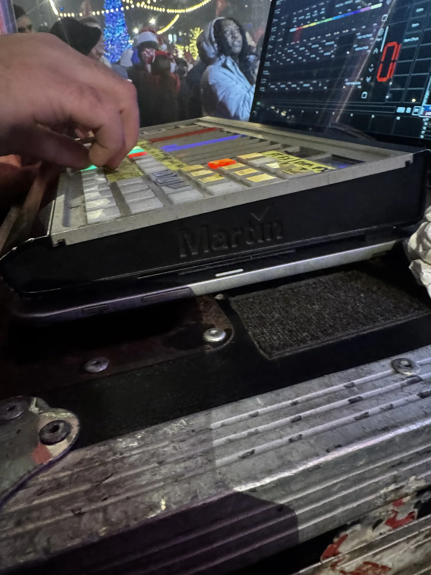 A hand on a Martin lighting console at the outdoor control position, crowd behind