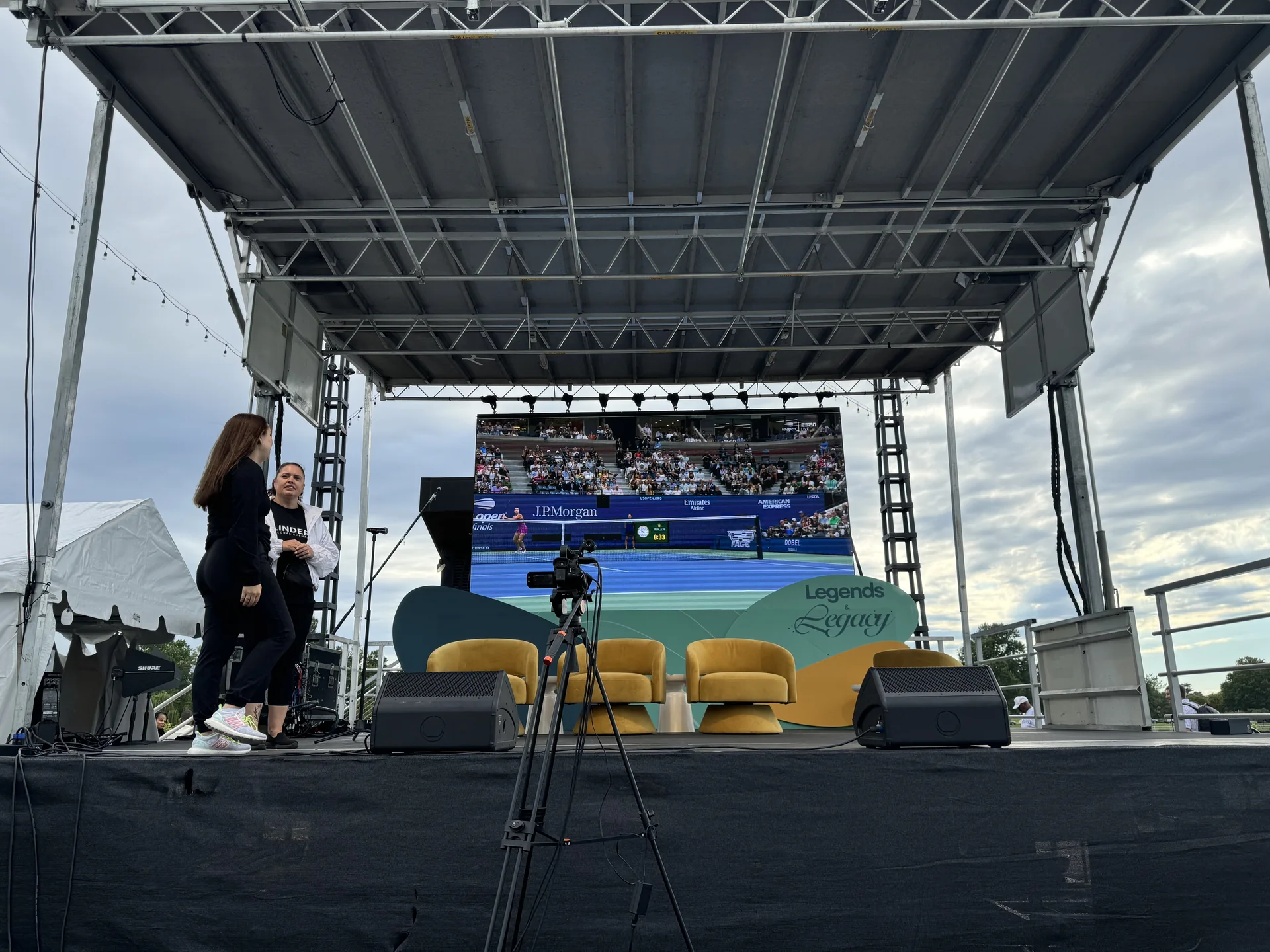 A camera on a tripod facing the stage screen showing the live broadcast