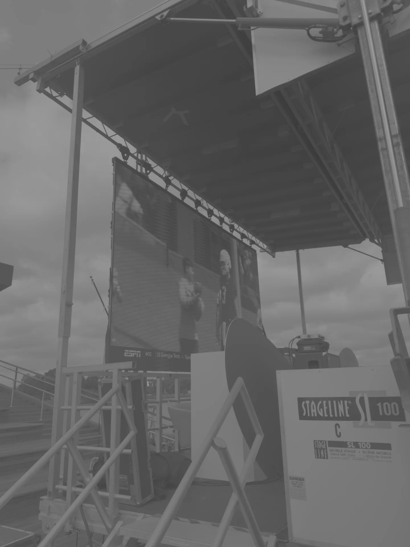 A tall screen tower seen from below running sports content, cloudy sky behind